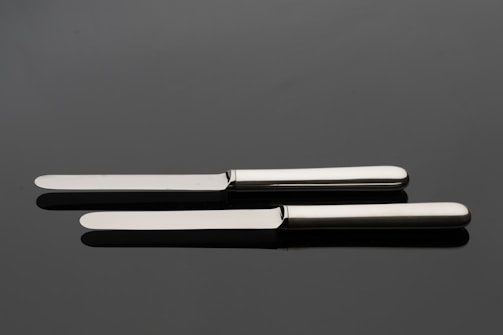 A pair of silver butter knives positioned parallel on a reflective dark surface.