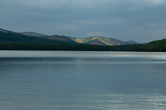 A serene landscape featuring a calm lake with gentle ripples on the surface. In the distance, lush forests line the shore, backed by rolling hills partially illuminated by sunlight breaking through a cloudy sky.