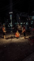 Construction workers are engaged in roadwork at night, illuminated by artificial lighting. They appear to be digging or inspecting a trench. The scene includes a background with construction equipment and a sign above that reads 'COFFEE'. The atmosphere suggests an urban setting with minimal traffic.