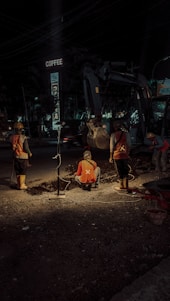 Construction workers are engaged in roadwork at night, illuminated by artificial lighting. They appear to be digging or inspecting a trench. The scene includes a background with construction equipment and a sign above that reads 'COFFEE'. The atmosphere suggests an urban setting with minimal traffic.
