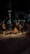 Construction workers are engaged in roadwork at night, illuminated by artificial lighting. They appear to be digging or inspecting a trench. The scene includes a background with construction equipment and a sign above that reads 'COFFEE'. The atmosphere suggests an urban setting with minimal traffic.