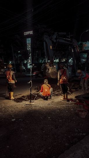 Construction workers are engaged in roadwork at night, illuminated by artificial lighting. They appear to be digging or inspecting a trench. The scene includes a background with construction equipment and a sign above that reads 'COFFEE'. The atmosphere suggests an urban setting with minimal traffic.