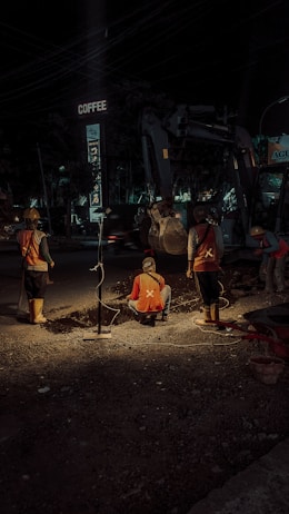Construction workers are engaged in roadwork at night, illuminated by artificial lighting. They appear to be digging or inspecting a trench. The scene includes a background with construction equipment and a sign above that reads 'COFFEE'. The atmosphere suggests an urban setting with minimal traffic.