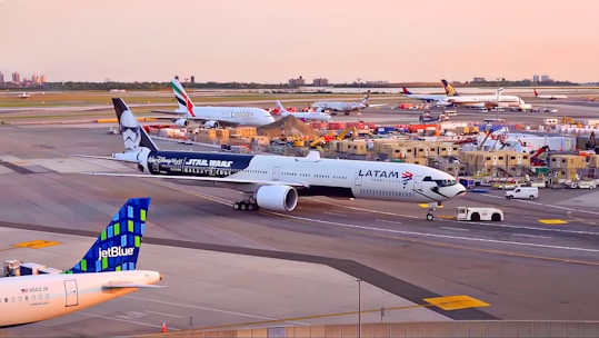 Several airplanes are positioned at an airport runway during sunset. The LATAM aircraft is prominently featured with a Star Wars-themed exterior. In the background, there are aircraft from Emirates and other airlines. Ground service vehicles and personnel can be seen alongside the small airport buildings.