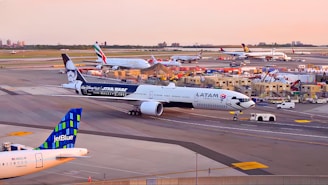 Several airplanes are positioned at an airport runway during sunset. The LATAM aircraft is prominently featured with a Star Wars-themed exterior. In the background, there are aircraft from Emirates and other airlines. Ground service vehicles and personnel can be seen alongside the small airport buildings.