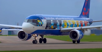 A commercial airplane features a vibrant paint job depicting cartoon characters along its fuselage, including blue humanoid figures with white hats and pants, possibly depicting characters from a well-known animated series. The aircraft is situated on an airport runway with green grass and a terminal visible in the background.