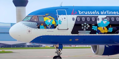 A Brussels Airlines airplane is parked at an airport, featuring animated Smurf characters on its exterior. One Smurf is dressed as a pilot, holding the controls in the cockpit, while another is dressed as a chef inside the plane.