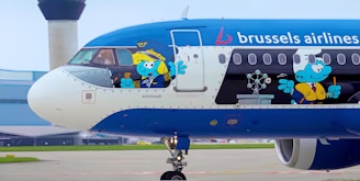 A Brussels Airlines airplane is parked at an airport, featuring animated Smurf characters on its exterior. One Smurf is dressed as a pilot, holding the controls in the cockpit, while another is dressed as a chef inside the plane.