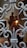 A close-up view of an intricately designed metal gate with ornamental patterns in the foreground. Behind the gate, there is a blurred statue of two figures, one larger and sitting and the other smaller and standing, possibly holding a lantern. The setting appears to be indoors, with arched doorways and a red light visible in the background.