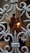 A close-up view of an intricately designed metal gate with ornamental patterns in the foreground. Behind the gate, there is a blurred statue of two figures, one larger and sitting and the other smaller and standing, possibly holding a lantern. The setting appears to be indoors, with arched doorways and a red light visible in the background.