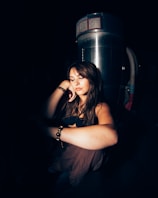 A woman is sitting in a dimly lit environment with a reflective metal structure behind her. She appears contemplative, resting her head on her hand. Her expression is serene and introspective.