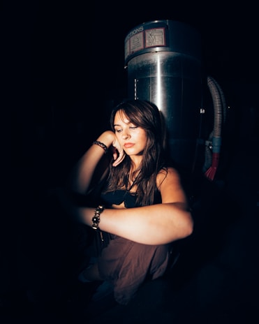 A woman is sitting in a dimly lit environment with a reflective metal structure behind her. She appears contemplative, resting her head on her hand. Her expression is serene and introspective.