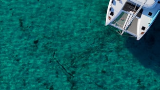An aerial view of a catamaran sailing on clear turquoise water. The boat is positioned towards the right of the image, with its white surfaces contrasting against the deep colors of the ocean. Shadows from the sail and frame create patterns on the deck.