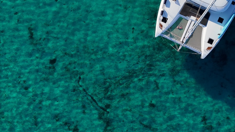 An aerial view of a catamaran sailing on clear turquoise water. The boat is positioned towards the right of the image, with its white surfaces contrasting against the deep colors of the ocean. Shadows from the sail and frame create patterns on the deck.