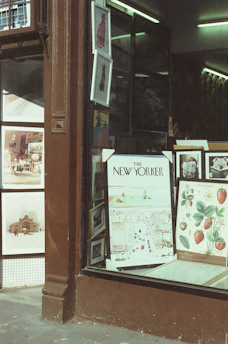 A storefront displays framed artwork and posters, including a prominent New Yorker cover and botanical illustrations of strawberries. The window is framed by a dark brown wood facade, and the interior is softly lit with fluorescent lights.