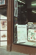 A storefront displays framed artwork and posters, including a prominent New Yorker cover and botanical illustrations of strawberries. The window is framed by a dark brown wood facade, and the interior is softly lit with fluorescent lights.