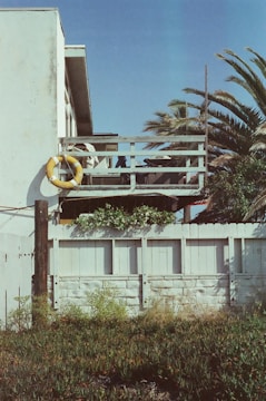 A white building with a balcony surrounded by a wooden fence. A yellow life preserver is hanging on the railing, and there are palm trees in the background.