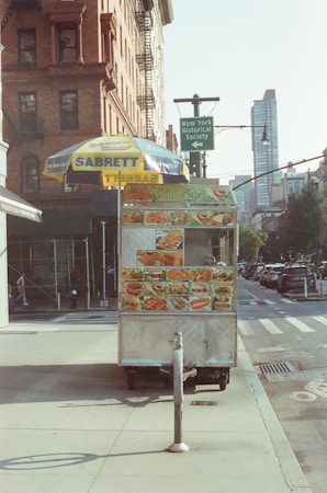 A street scene featuring a food cart with colorful images of various dishes. The cart is located on a sidewalk next to a building, topped with an umbrella bearing the brand 'Sabrett'. There is a sign indicating the nearby New York Historical Society, and a street lined with parked cars and tall buildings is visible in the background.