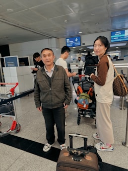 A busy airport setting with multiple people in the scene. Two people are in the foreground, one carrying a bag and the other standing near a suitcase. Other travelers can be seen in the background near a check-in counter, and there are signs displaying flight information overhead.
