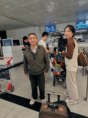 A busy airport setting with multiple people in the scene. Two people are in the foreground, one carrying a bag and the other standing near a suitcase. Other travelers can be seen in the background near a check-in counter, and there are signs displaying flight information overhead.