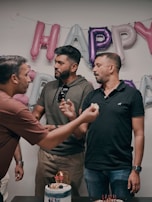 Three men are gathered in front of a wall with shiny inflated balloons that spell out a festive message. Two of the men are engaged in feeding each other cake, with one holding a piece of cake in his hand. A decorated cake with lit candles is placed on the table in front of them. They appear to be celebrating a special occasion or party in a casual setting.