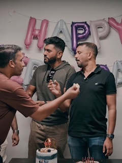 Three men are gathered in front of a wall with shiny inflated balloons that spell out a festive message. Two of the men are engaged in feeding each other cake, with one holding a piece of cake in his hand. A decorated cake with lit candles is placed on the table in front of them. They appear to be celebrating a special occasion or party in a casual setting.