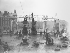 Workers are engaged in construction or maintenance on a bridge with large structures and visible machinery. The setting appears industrial, with several people actively working and smoke rising from various points. Buildings are visible in the background, and the bridge features signage with the phrase 'STOP FOR BUNK.'
