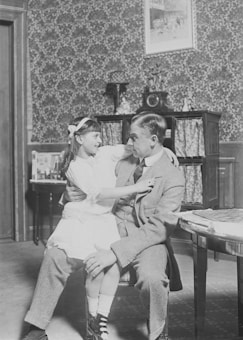 A young girl is sitting on the lap of an older man dressed in a suit. They appear to be in a vintage-style room with patterned wallpaper, a framed picture on the wall, and a cabinet with decorative items in the background. The girl is smiling at the man, and they both seem to be enjoying a pleasant moment together.