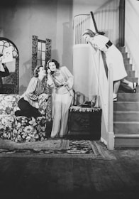 Three women are interacting in a domestic setting. Two women are seated on a floral patterned couch, sharing a laugh, while one is holding a telephone. Another woman leans over the stair railing, smiling down at them. The room features a patterned carpet, a staircase, and a window with patterned curtains.