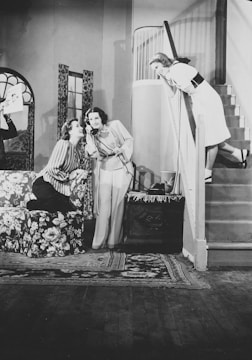 Three women are interacting in a domestic setting. Two women are seated on a floral patterned couch, sharing a laugh, while one is holding a telephone. Another woman leans over the stair railing, smiling down at them. The room features a patterned carpet, a staircase, and a window with patterned curtains.