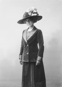 A woman in vintage attire is standing against a plain backdrop. She wears a dark velvet suit with a high-collared blouse underneath. Her ensemble includes a long skirt and a tailored jacket with decorative buttons. On her head, she has an elaborate wide-brimmed hat adorned with flowers, adding a touch of elegance. Her expression is calm and composed.