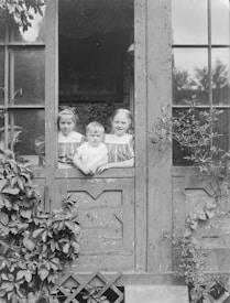 Three children are standing behind a wooden gate, with ivy growing on it. They are peering through a window with large glass panes. The children are dressed in light-colored clothing, with two girls on either side of a younger boy in the middle. The setting appears to be outdoors, with foliage visible through the window.