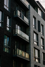 A modern apartment building with dark brick walls and large glass windows. The structure features several balconies with reflective glass railings. The building has an angular, geometric design, creating a striking contrast between the brick and concrete sections.