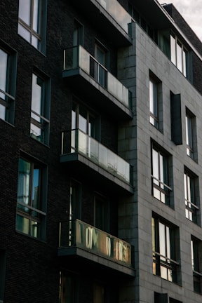 A modern apartment building with dark brick walls and large glass windows. The structure features several balconies with reflective glass railings. The building has an angular, geometric design, creating a striking contrast between the brick and concrete sections.