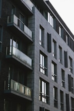 A modern apartment building with a minimalist design features large glass windows and balconies. The structure is composed of gray stone bricks and dark walls, giving it a contemporary and urban appearance.