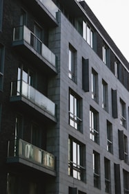 A modern apartment building with a minimalist design features large glass windows and balconies. The structure is composed of gray stone bricks and dark walls, giving it a contemporary and urban appearance.