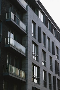 A modern apartment building with a minimalist design features large glass windows and balconies. The structure is composed of gray stone bricks and dark walls, giving it a contemporary and urban appearance.