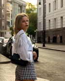A young woman with blond hair is walking along a city street. She is wearing a white blouse, a black and white houndstooth skirt, and is carrying a black handbag with a gold chain strap. The background features buildings, parked cars, and a street lamp with trees visible in the distant urban environment. The street appears quiet with few or no people around.
