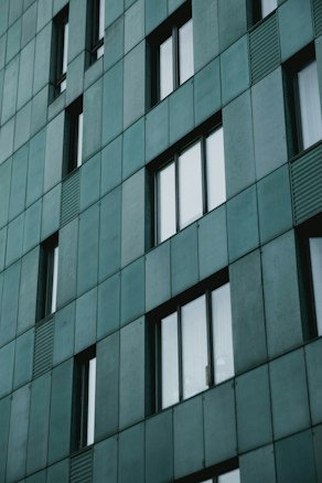 A modern building facade featuring a grid pattern of rectangular, teal-colored panels. The design includes vertical and horizontal lines with uniform windows slightly protruding from the surface, adding depth.