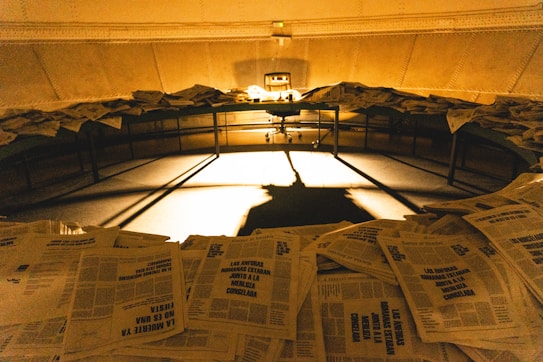 A dimly lit room filled with stacks of newspapers scattered across a circular table. Shadows are cast dramatically on the floor by the light in the center, creating a mysterious and somewhat chaotic ambiance.