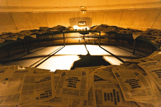 A dimly lit room filled with stacks of newspapers scattered across a circular table. Shadows are cast dramatically on the floor by the light in the center, creating a mysterious and somewhat chaotic ambiance.