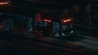 A bus is parked at a dimly lit bus station during nighttime. The bus has glowing red and white LED lights displaying route 69. The surrounding area is dark, with shadows cast on the ground and minimal ambient light. The station appears empty, with no visible people around.