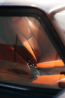 The image depicts the interior of a car viewed through a window, showcasing orange car seats with a modern design. The seats are illuminated by natural light, and the window reflects some external features.