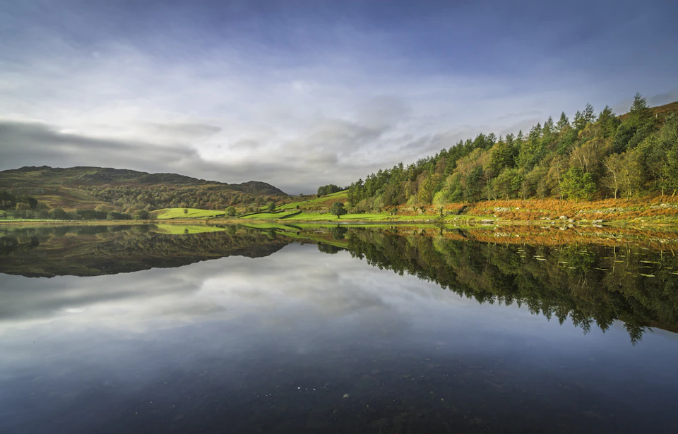 The image depicts a serene lake surrounded by lush green trees and rolling hills, creating a picturesque landscape. The calm water reflects the surrounding greenery, adding depth to the scene. The sky above is filled with clouds, casting a soft light over the entire area. The perspective of the photo is from the shoreline, looking out towards the horizon where the lake meets the land.