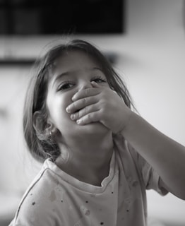 A young child with long hair is covering their mouth with one hand and looking directly at the camera. They are wearing a light-colored shirt with small designs on it. The background is blurry and features a dark rectangular shape, possibly a television.