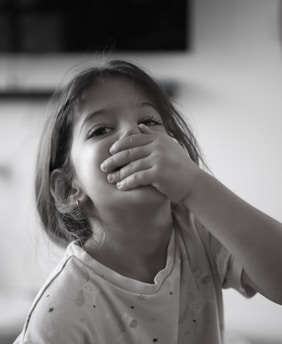 A young child with long hair is covering their mouth with one hand and looking directly at the camera. They are wearing a light-colored shirt with small designs on it. The background is blurry and features a dark rectangular shape, possibly a television.