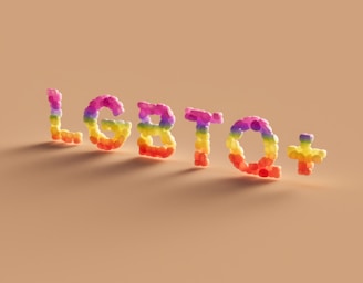 The image features the letters 'LGBTQ+' formed by colorful balloon-like structures on a light brown background. Each letter is constructed from balloons of various colors, including pink, purple, orange, yellow, green, and red.