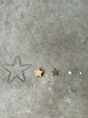 A collection of five star shapes of varying sizes lined up on a concrete surface. The stars range from a large wireframe outline to small solid forms, with one being golden and others in shades of brown, silver, and white.