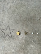 A collection of five star shapes of varying sizes lined up on a concrete surface. The stars range from a large wireframe outline to small solid forms, with one being golden and others in shades of brown, silver, and white.