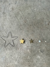 A collection of five star shapes of varying sizes lined up on a concrete surface. The stars range from a large wireframe outline to small solid forms, with one being golden and others in shades of brown, silver, and white.